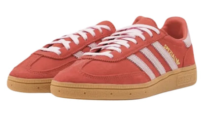 Een paar Adidas Handball Spezial Bright Red/Clear Pink sneakers met witte strepen, witte veters en een lichtbruine rubberen zool. Op de zijkant van de schoenen staat het woord "SPEZIAL" in gouden tekst, wat doet denken aan de Adidas Handball Spezial-stijl.