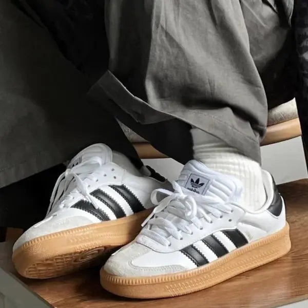 Close-up van een persoon die witte Adidas Samba XLG wit/zwarte sneakers draagt met zwarte strepen en een rubberen zool, gecombineerd met witte geribbelde sokken en een grijze broek.
